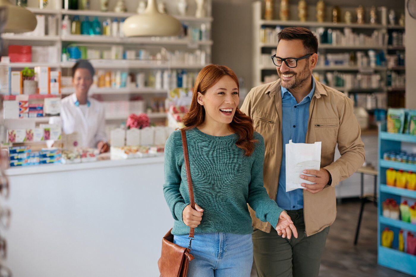 A couple leaving a pharmacy with their prescription, pharmacist smiling behind the counter